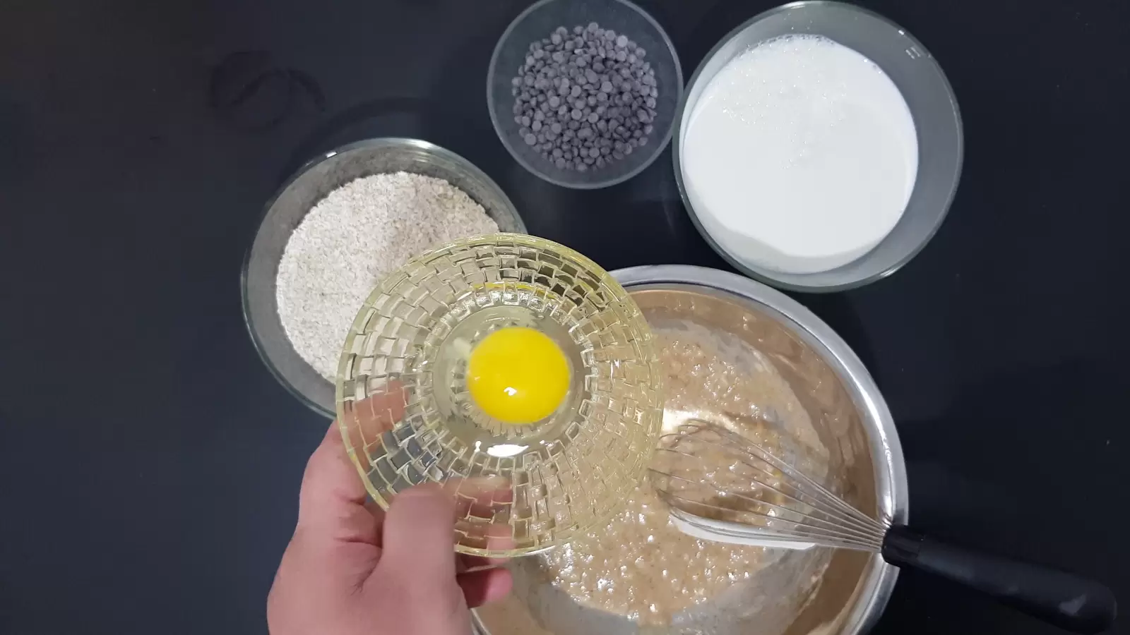 Bowl with chocolate chips, bowl with egg, bowl with oats, bowl with milk, whisk, mixture in a bowl