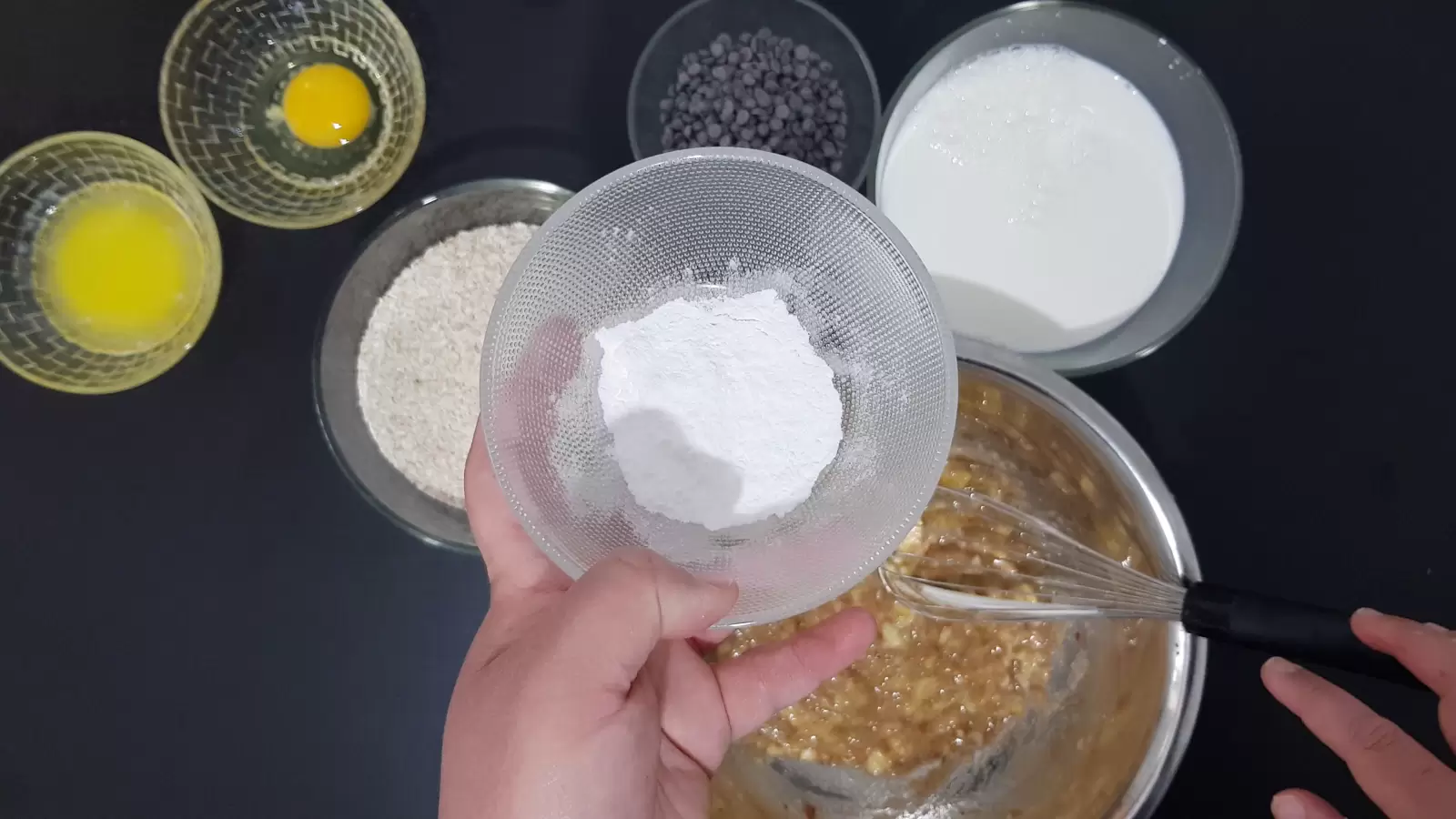 Bowl with baking powder, bowl with chocolate chips, bowl with egg, bowl with butter, bowl with oats, bowl with milk, whisk, mashed bananas in a bowl