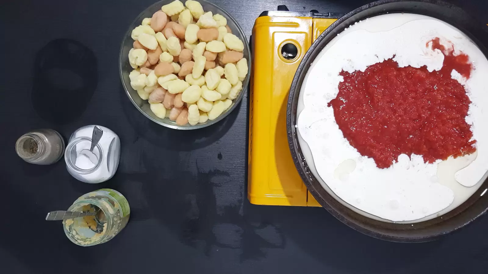 a frying pan with whipping cream and crushed tomatoes, a bowl of gnocchi, a jar of salt and a spoon, a jar of crushed garlic, a jar of black pepper
