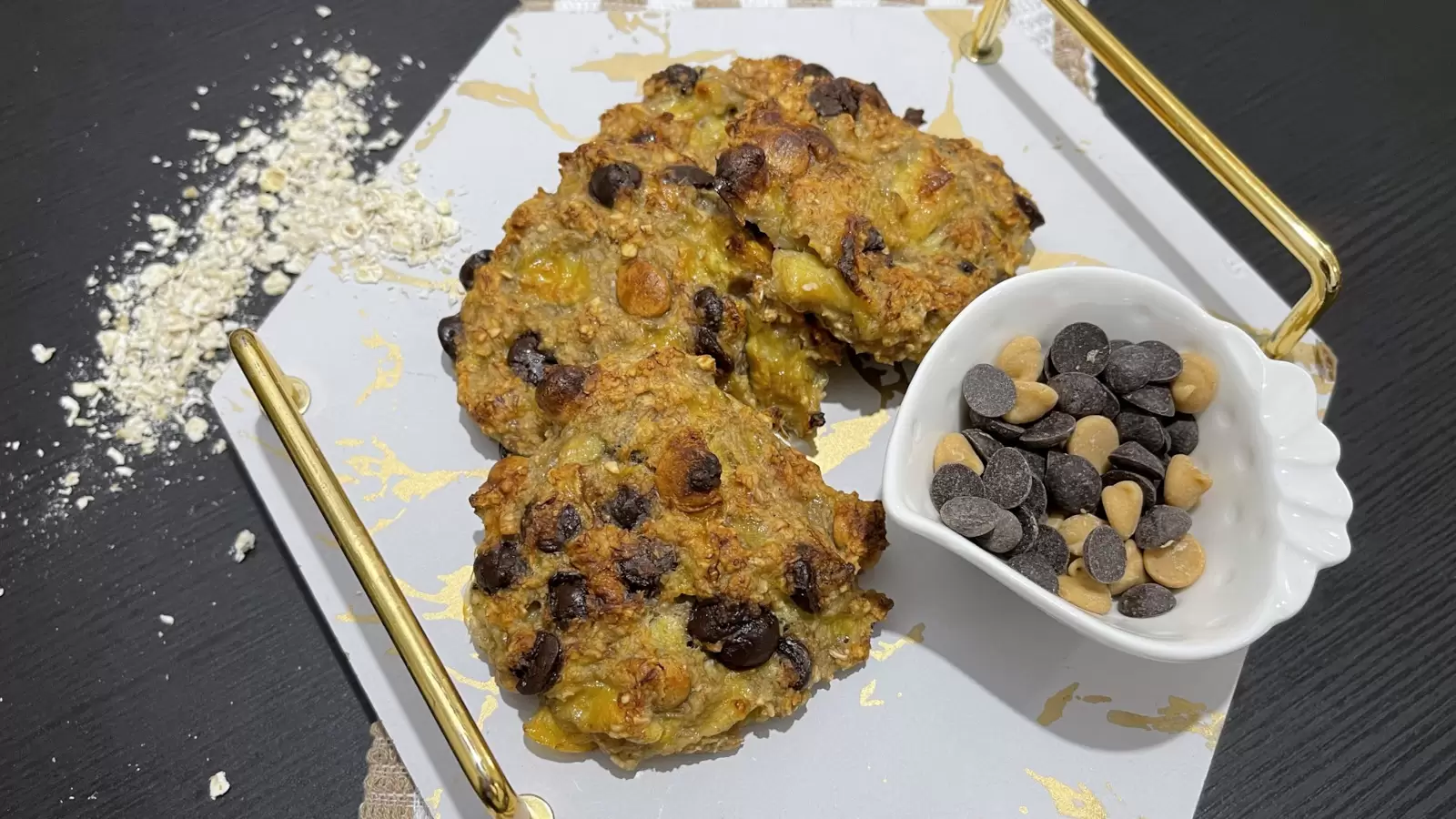 Banana and oatmeal cookies on a tray a bowl of chocolate chips oatmeal scattered on the table