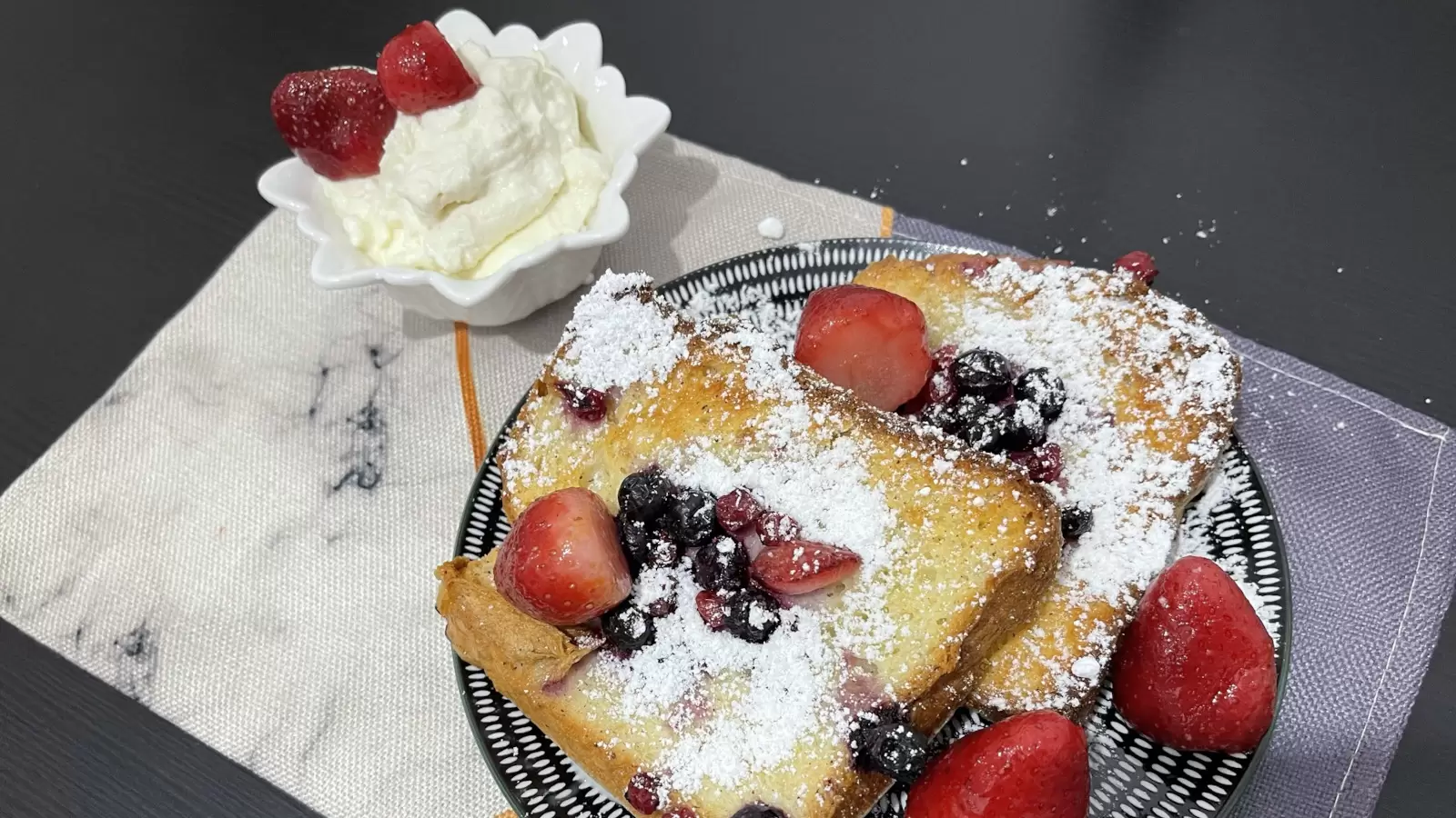 Recipe for Berry Bread Pudding on a plate next to vanilla cream with strawberries in a bowl on a table