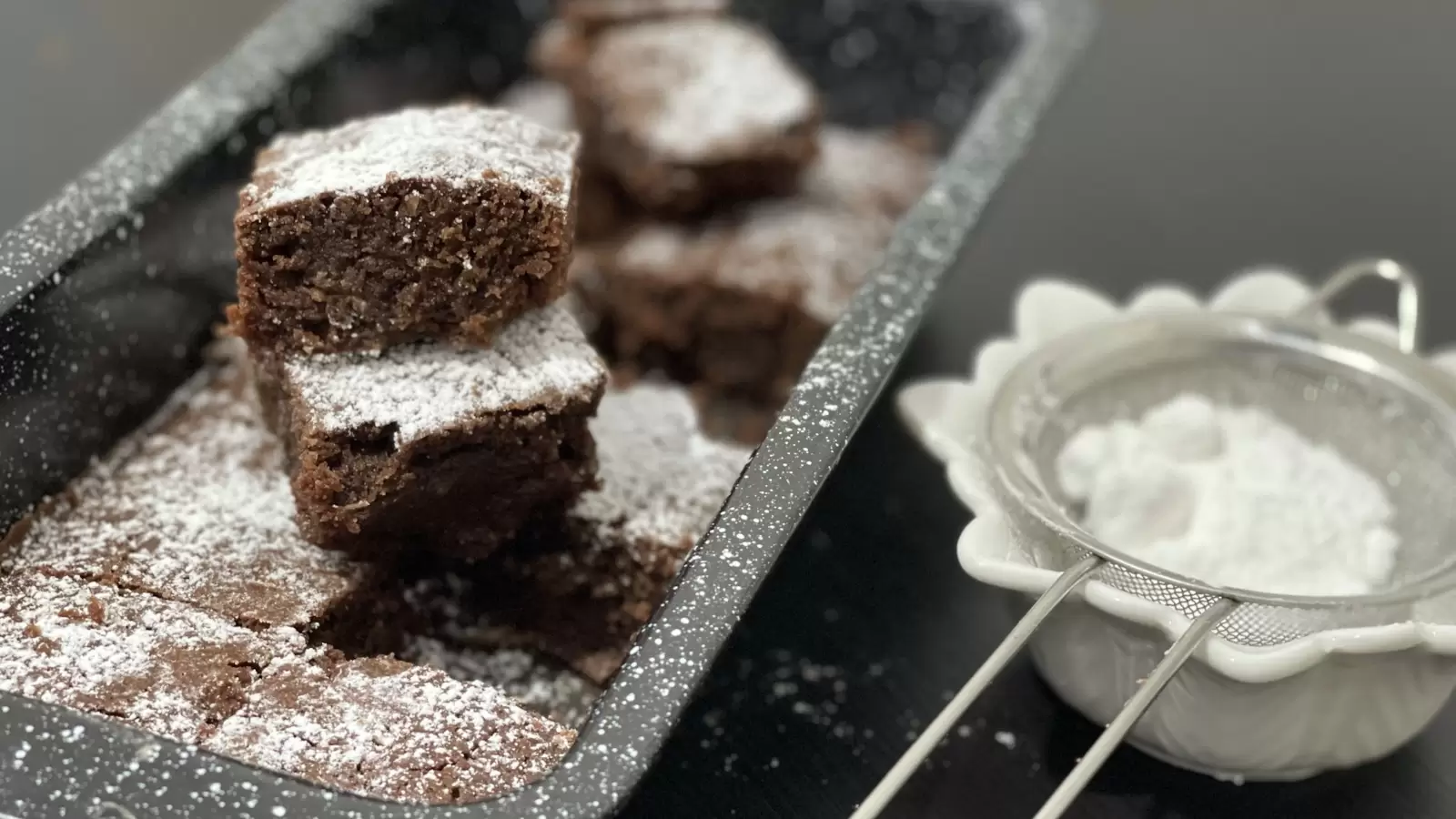 Brownies in a pan brownie pieces one on top of the other next to powdered sugar in a bowl