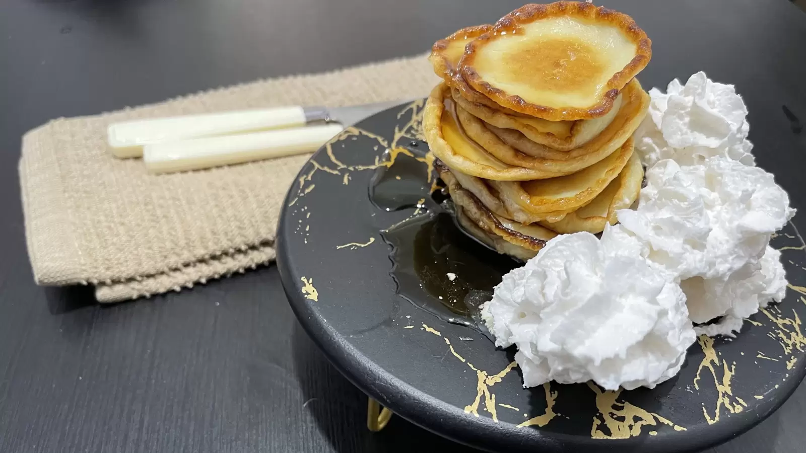 Cheese pancakes whipped cream on a black surface next to a towel with a fork and knife on a black table