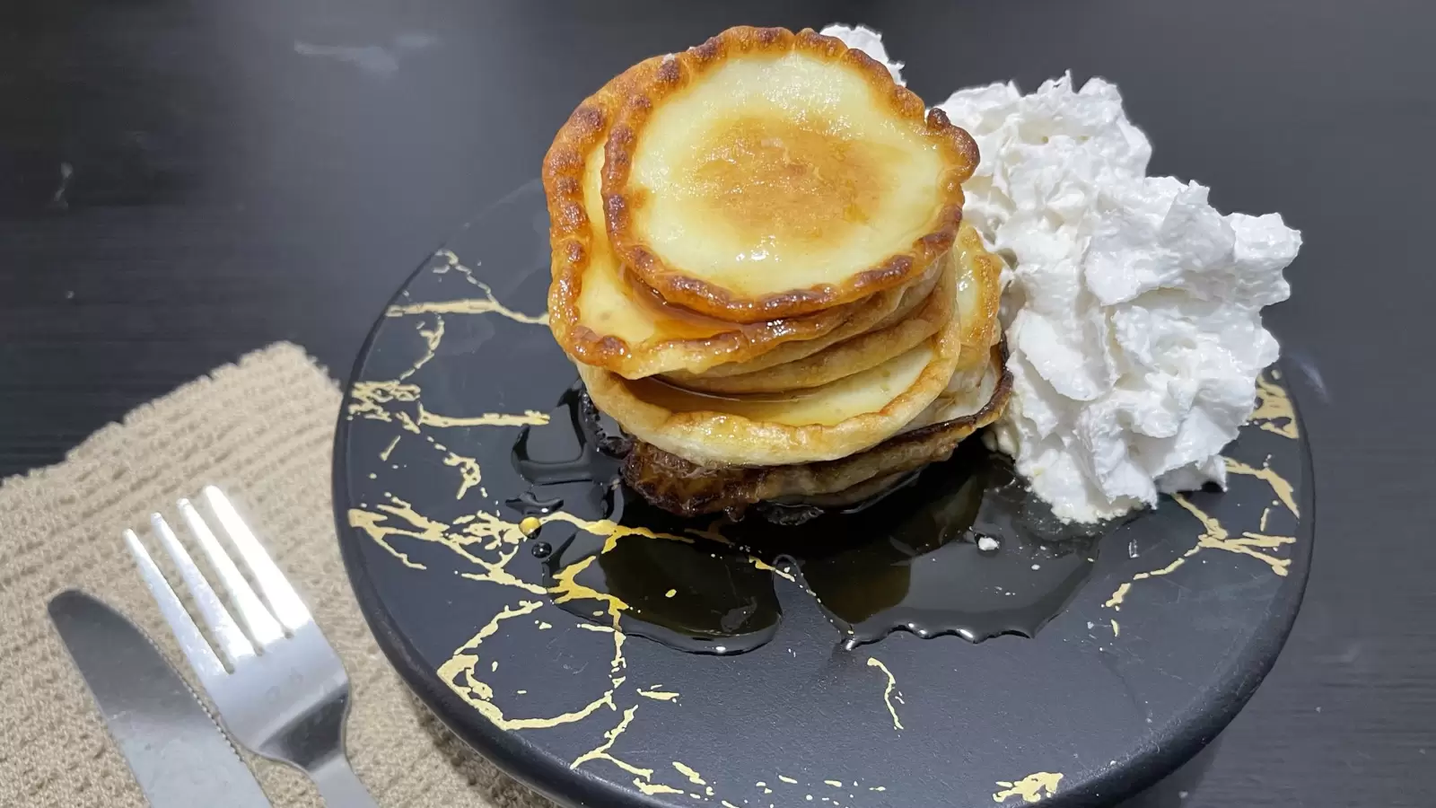 Cheese pancakes recipe whipped cream on a black surface next to a towel with a fork and knife on a black table