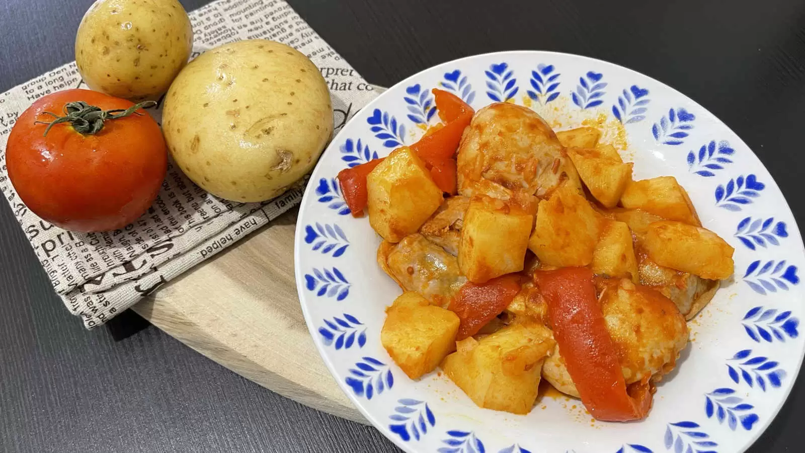 A plate with a dish of chicken potatoes in tomato sauce a wooden board on it a napkin two potatoes and one tomato