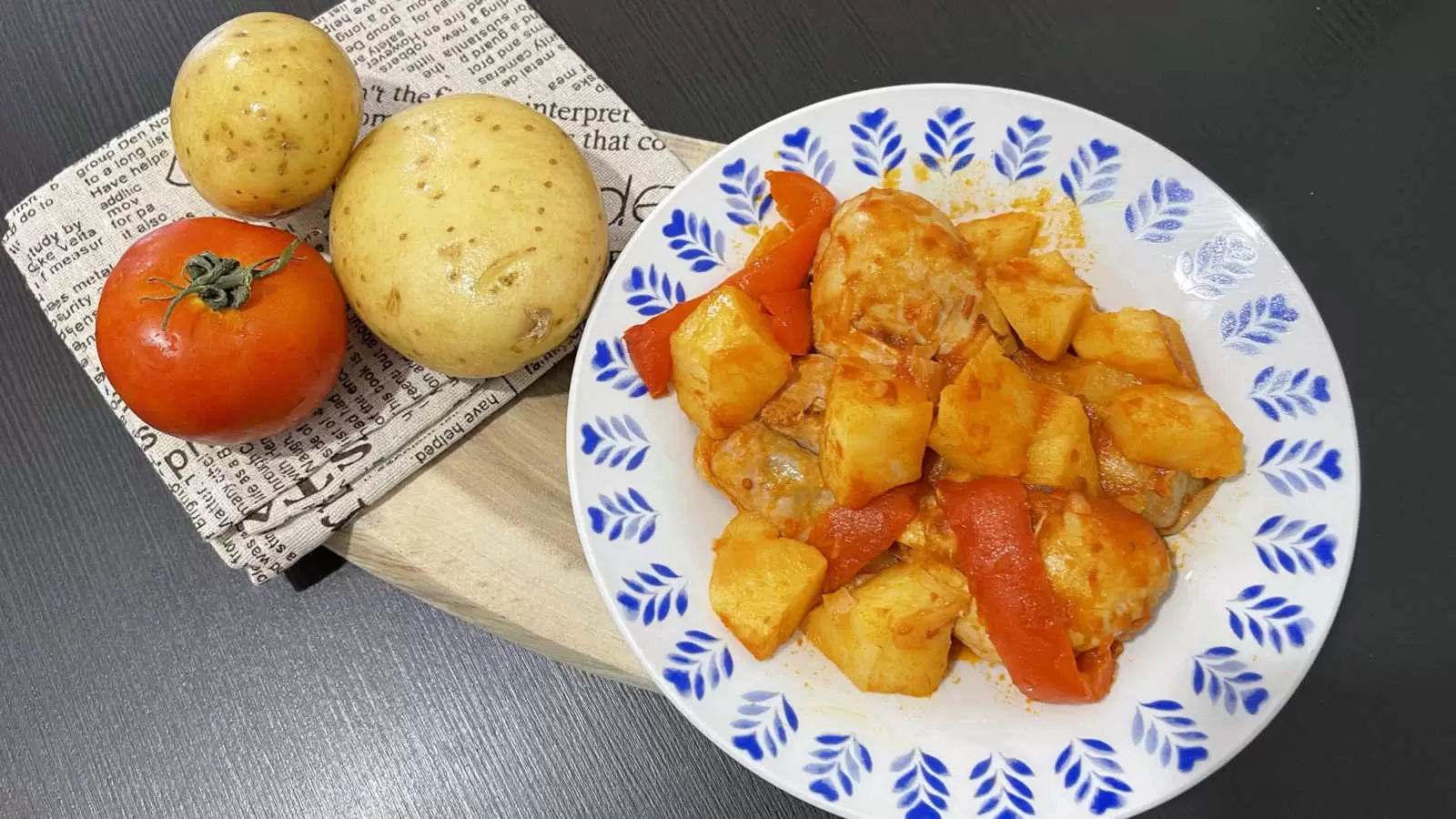 A plate with a dish of chicken potatoes in tomato sauce a wooden board on it a napkin two potatoes and one tomato