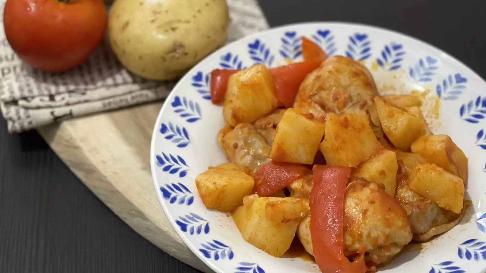 A plate with a meal of chicken potatoes in tomato sauce a wooden board on it a napkin potato and tomato