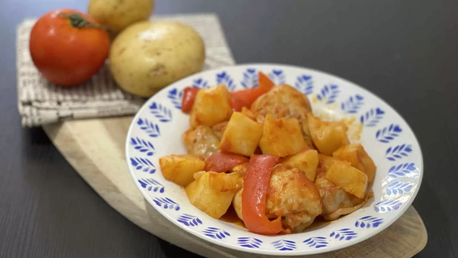 A plate with a dish of chicken potatoes in tomato sauce in one pot a wooden board on it a napkin two potatoes and one tomato