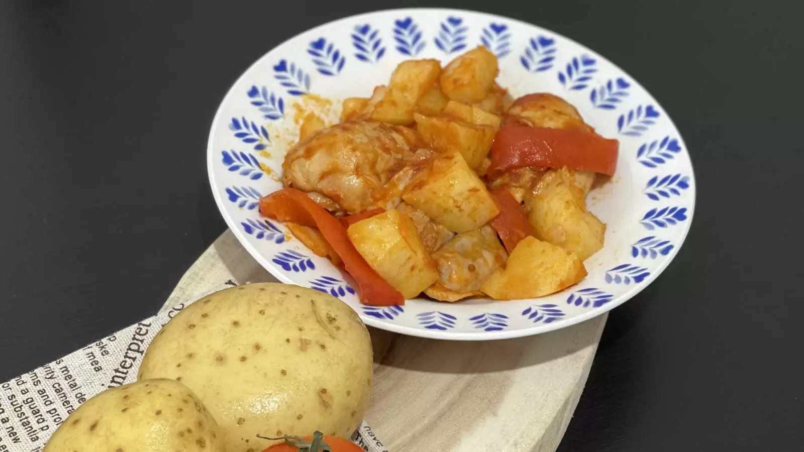 A plate with a dish of chicken potatoes in tomato sauce recipe a wooden board on it a napkin two potatoes and one tomato