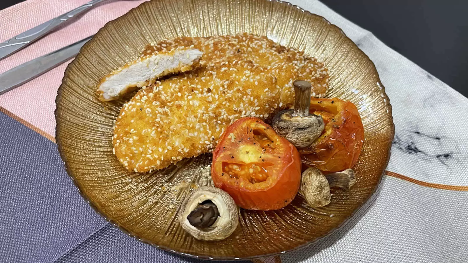 Chicken Schnitzel roasted tomatoes and mushrooms on a gold colored plate on a napkin and a fork and knife on the side