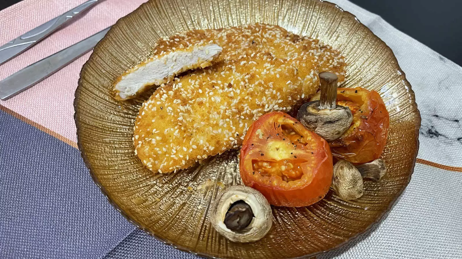 Chicken Schnitzel dish roasted tomatoes and mushrooms on a gold colored plate on a napkin and a fork and knife on the side