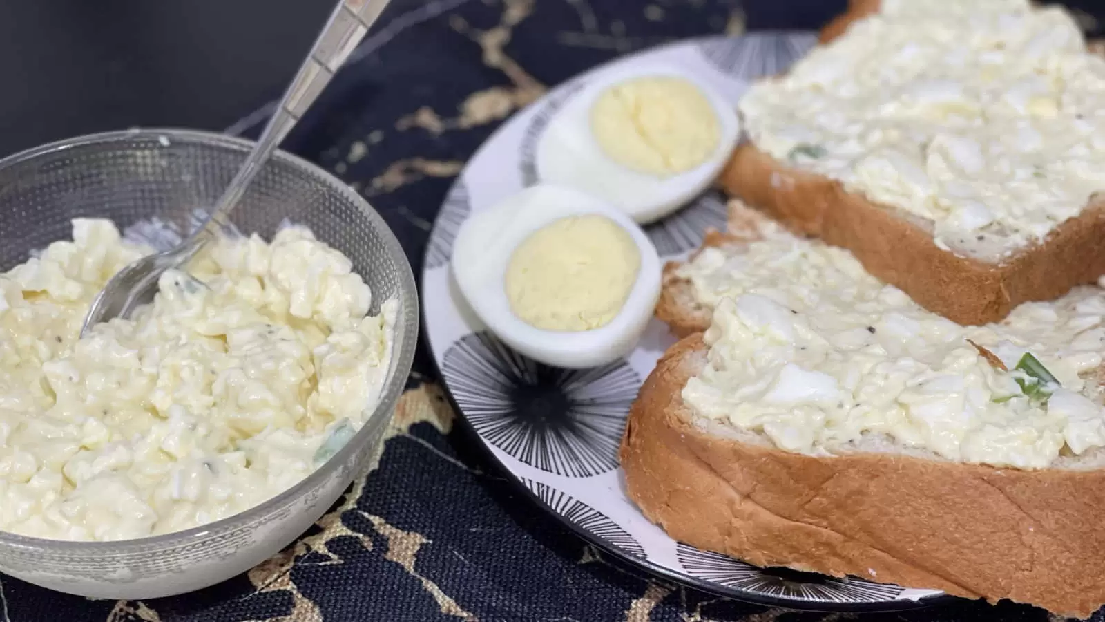 A bowl with egg salad and a spoon a plate with two slices of bread with egg salad spread a hard boiled egg cut in half