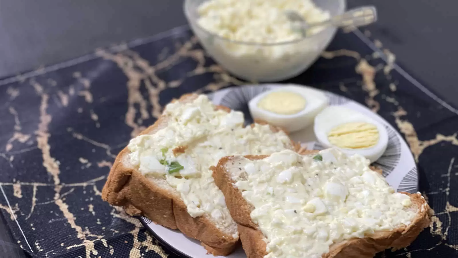 A bowl with egg salad dish and a spoon a plate with two slices of bread with egg salad spread a hard boiled egg cut in half