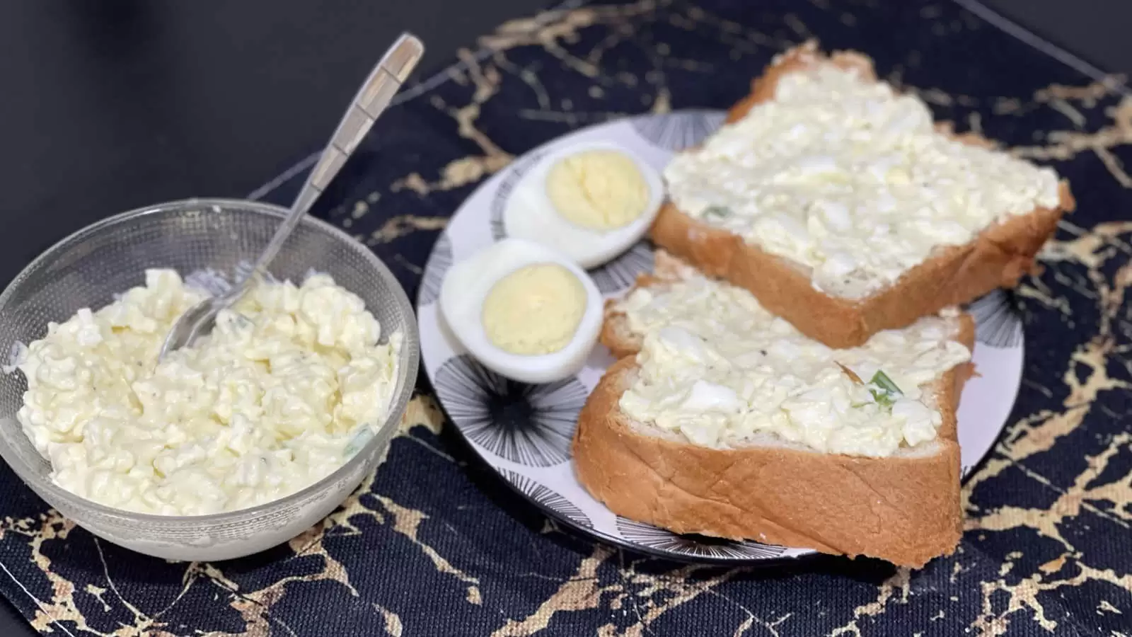A bowl with egg salad recipe and a spoon a plate with two slices of bread with egg salad spread a hard boiled egg cut in half