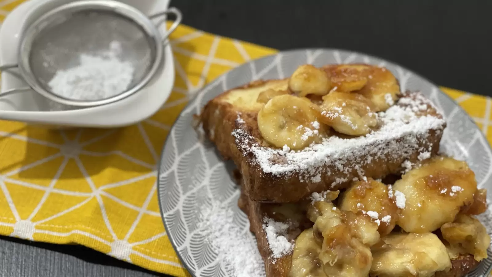 French toast and caramelized bananas on a plate powdered sugar in a bowl next to it