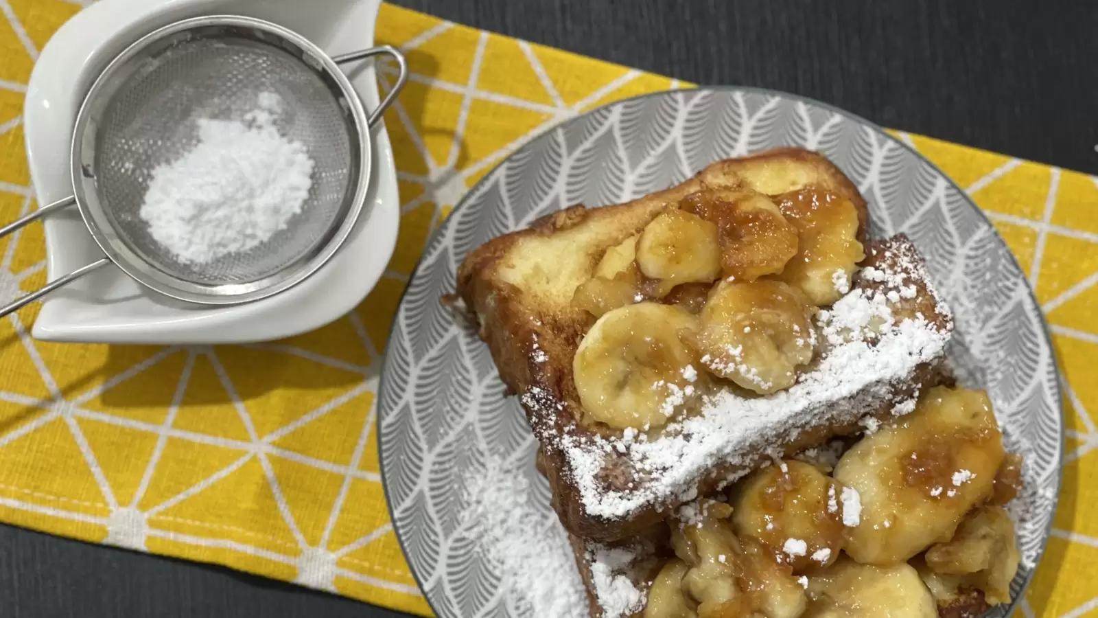 French toast and caramelized bananas dish on a plate powdered sugar in a bowl next to it