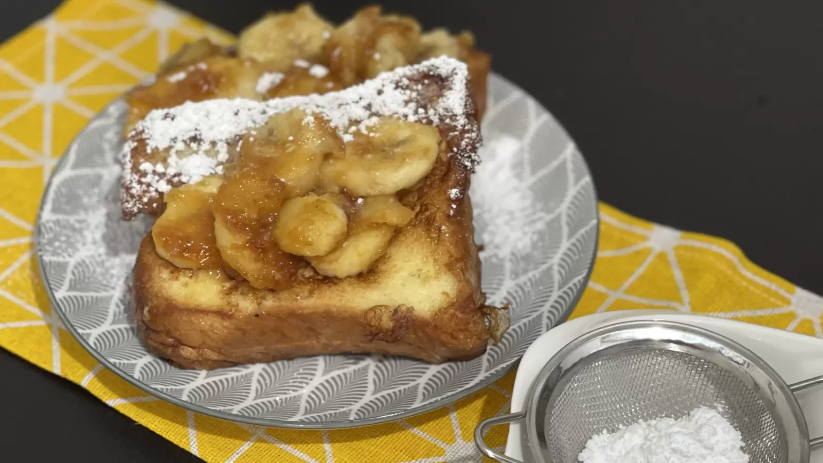 French toast and caramelized bananas recipe on a plate powdered sugar in a bowl next to it