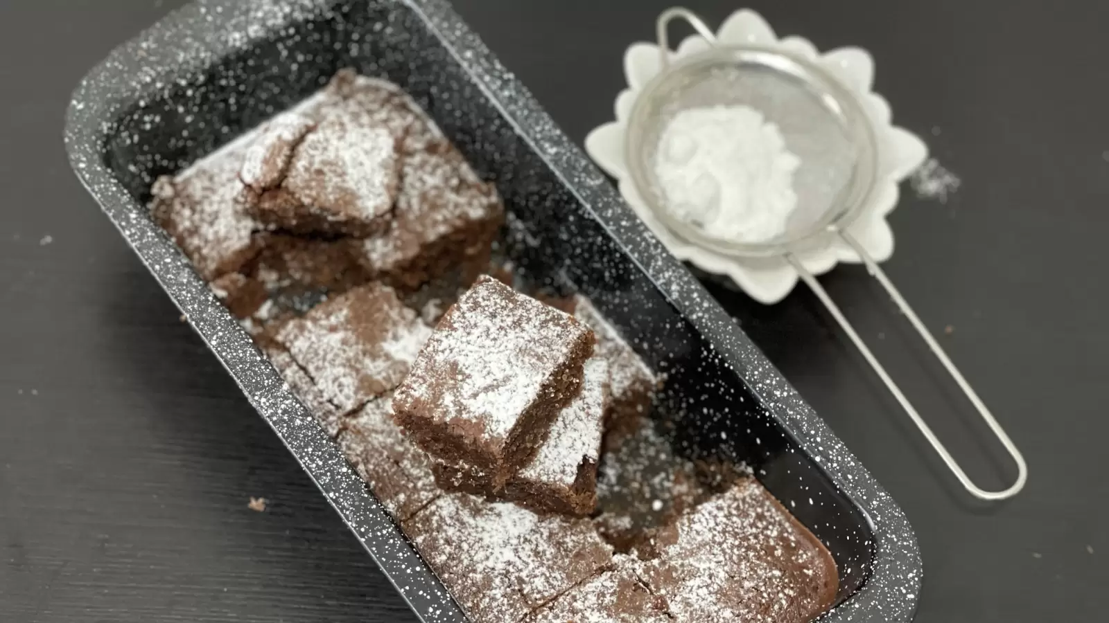 Homemade Brownies in a pan brownie pieces one on top of the other next to powdered sugar in a bowl