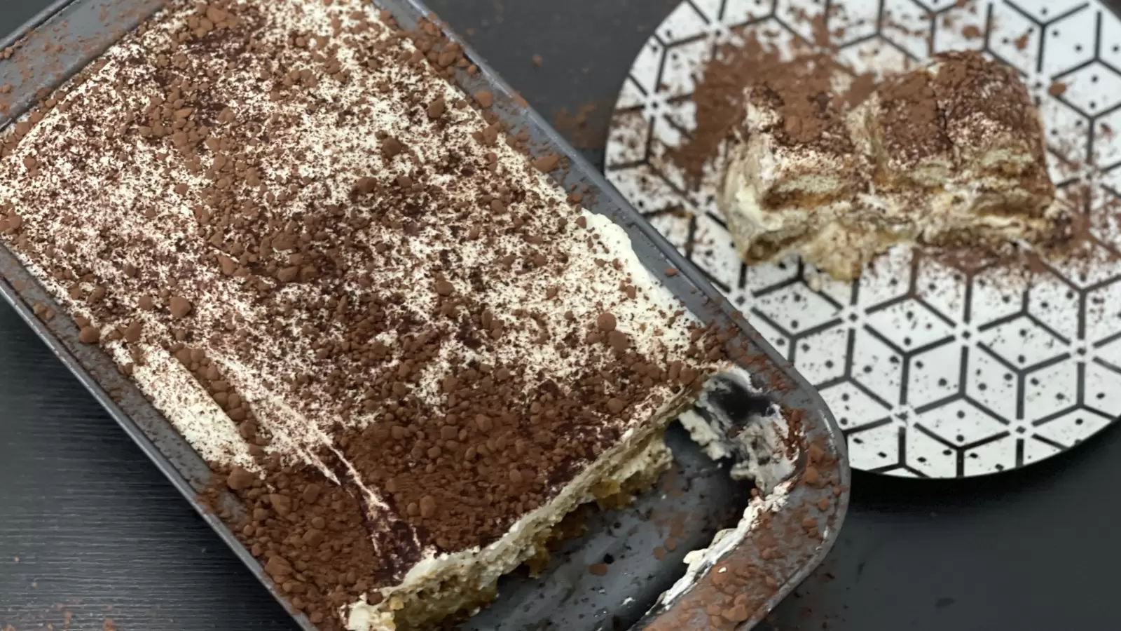 A piece of Homemade tiramisu on a plate and a tiramisu cake in a pan next to it