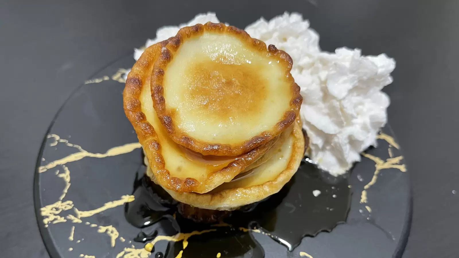 Homemade Cheese pancakes whipped cream on a black surface next to a towel with a fork and knife on a black table