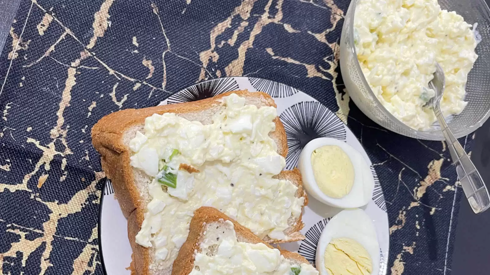 A bowl with homemade egg salad and a spoon a plate with two slices of bread with egg salad spread a hard boiled egg cut in half