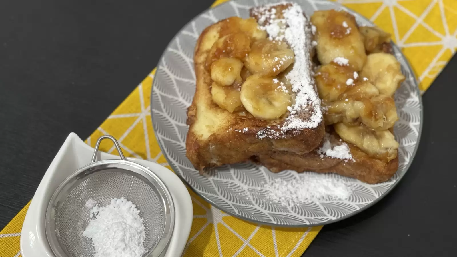 Homemade French toast and caramelized bananas on a plate powdered sugar in a bowl next to it
