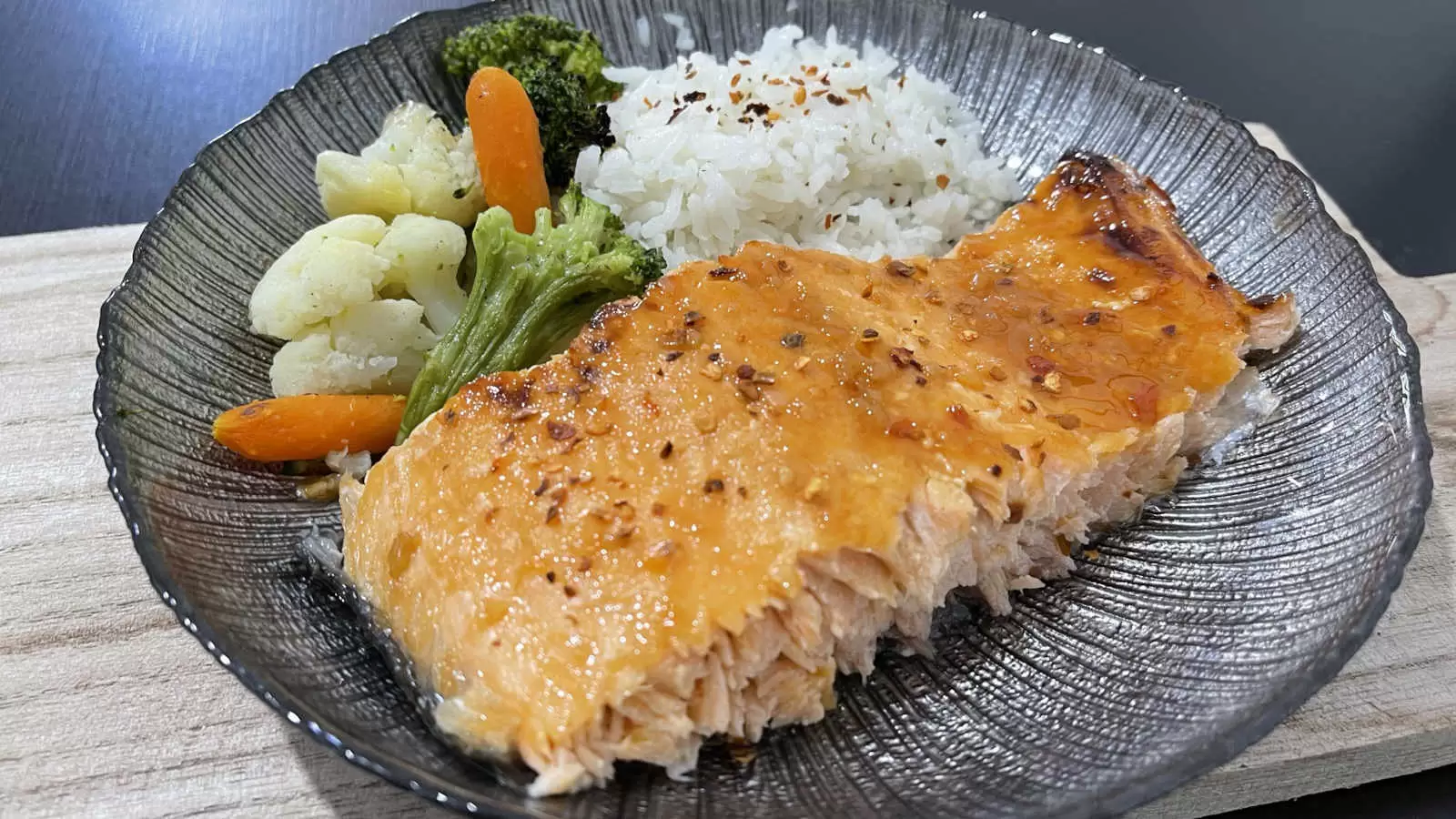 Honey Glazed Salmon rice carrots broccoli and cauliflower on a black plate on a wooden board