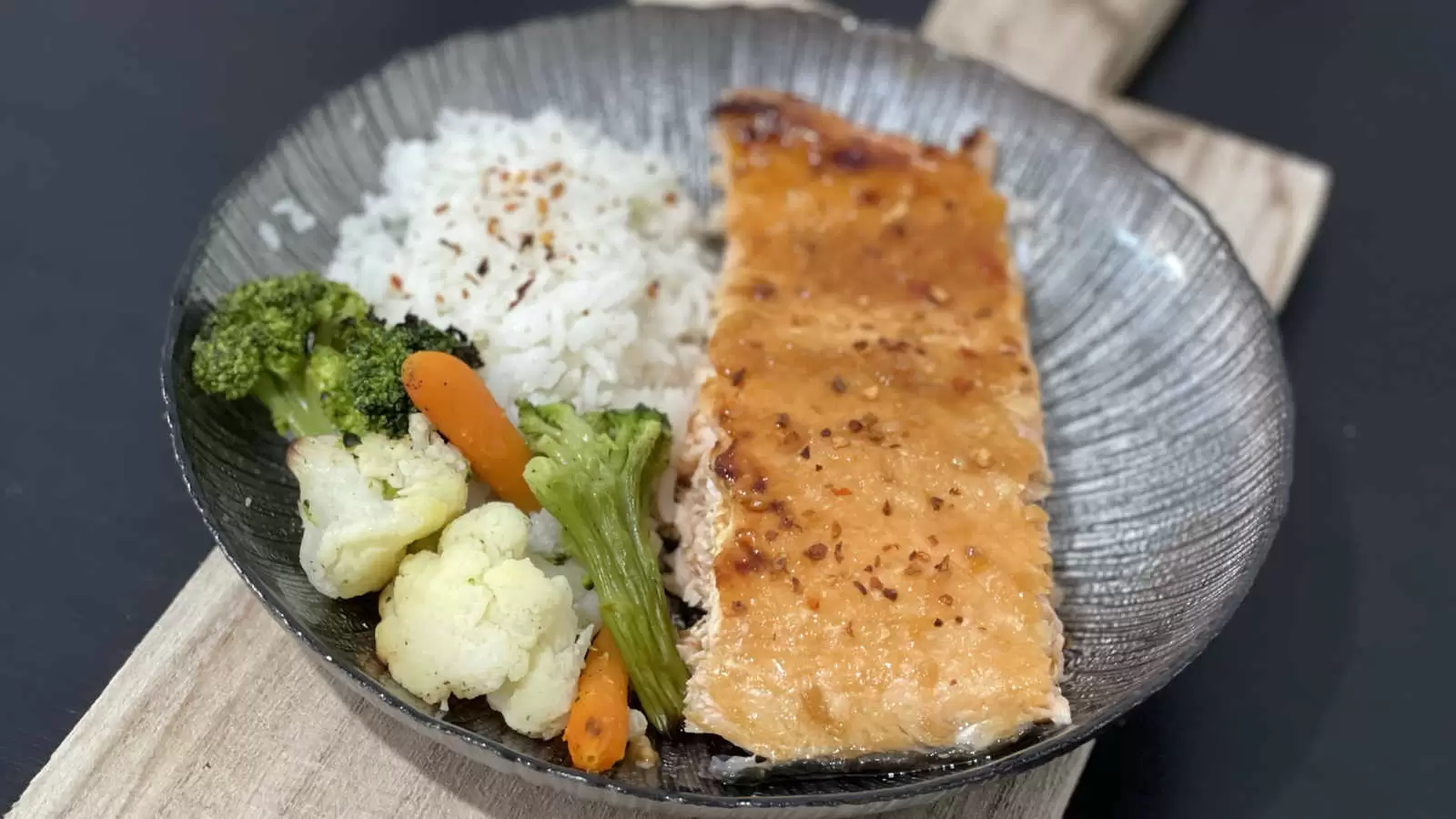 Honey Glazed Salmon dish with rice carrots broccoli and cauliflower on a black plate on a wooden board