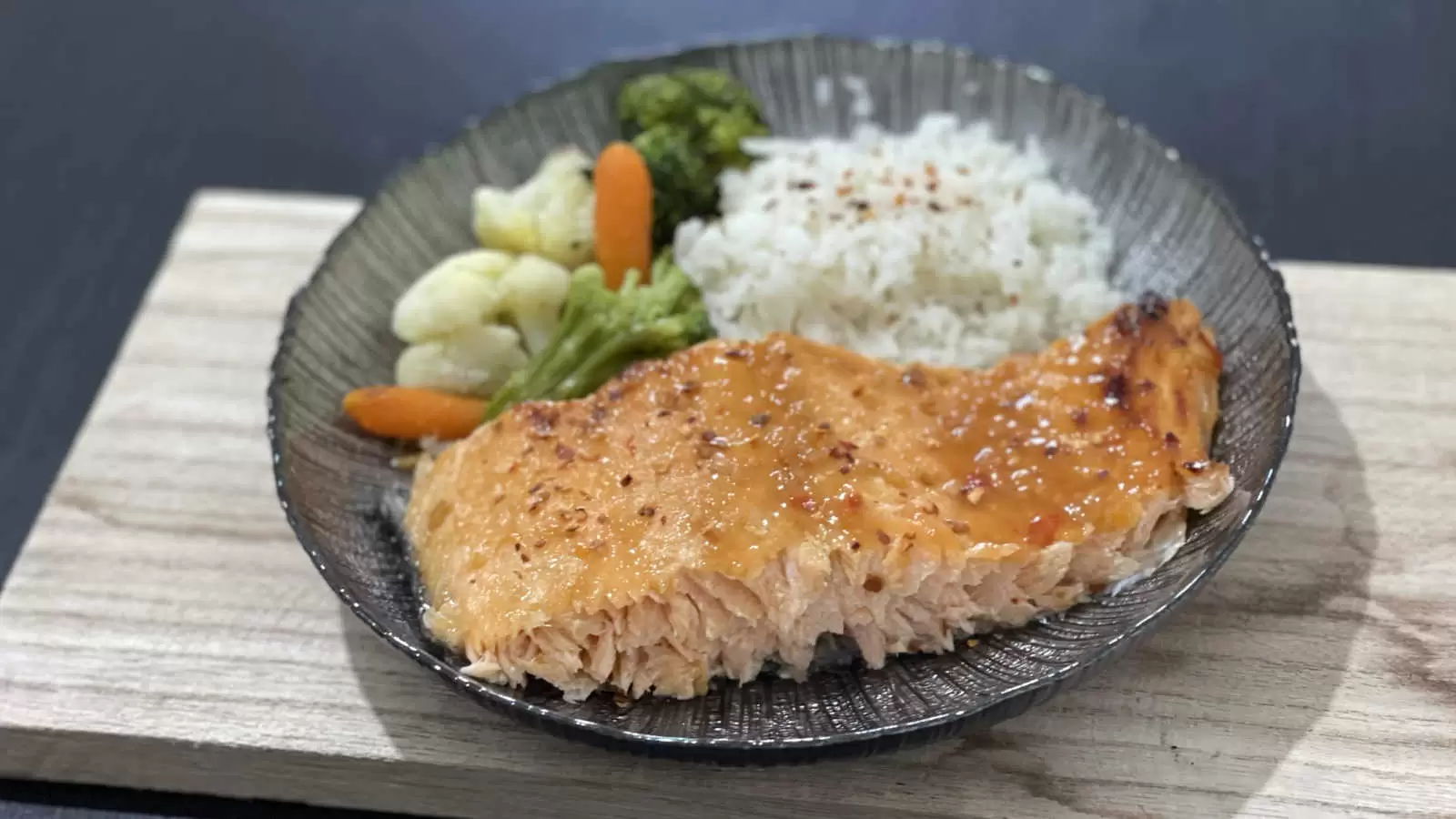 Honey Glazed Salmon Recipe with rice carrots broccoli and cauliflower on a black plate on a wooden board