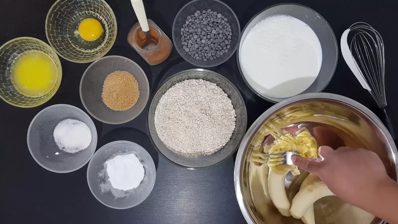 Bowl with baking powder, bowl with chocolate chips, cinnamon and spoon, bowl with egg, bowl with butter, bowl with brown sugar, bowl with oats, bowl with milk, bowl with vanilla sugar, bowl with baking powder, fork, whisk , mash bananas over a bowl