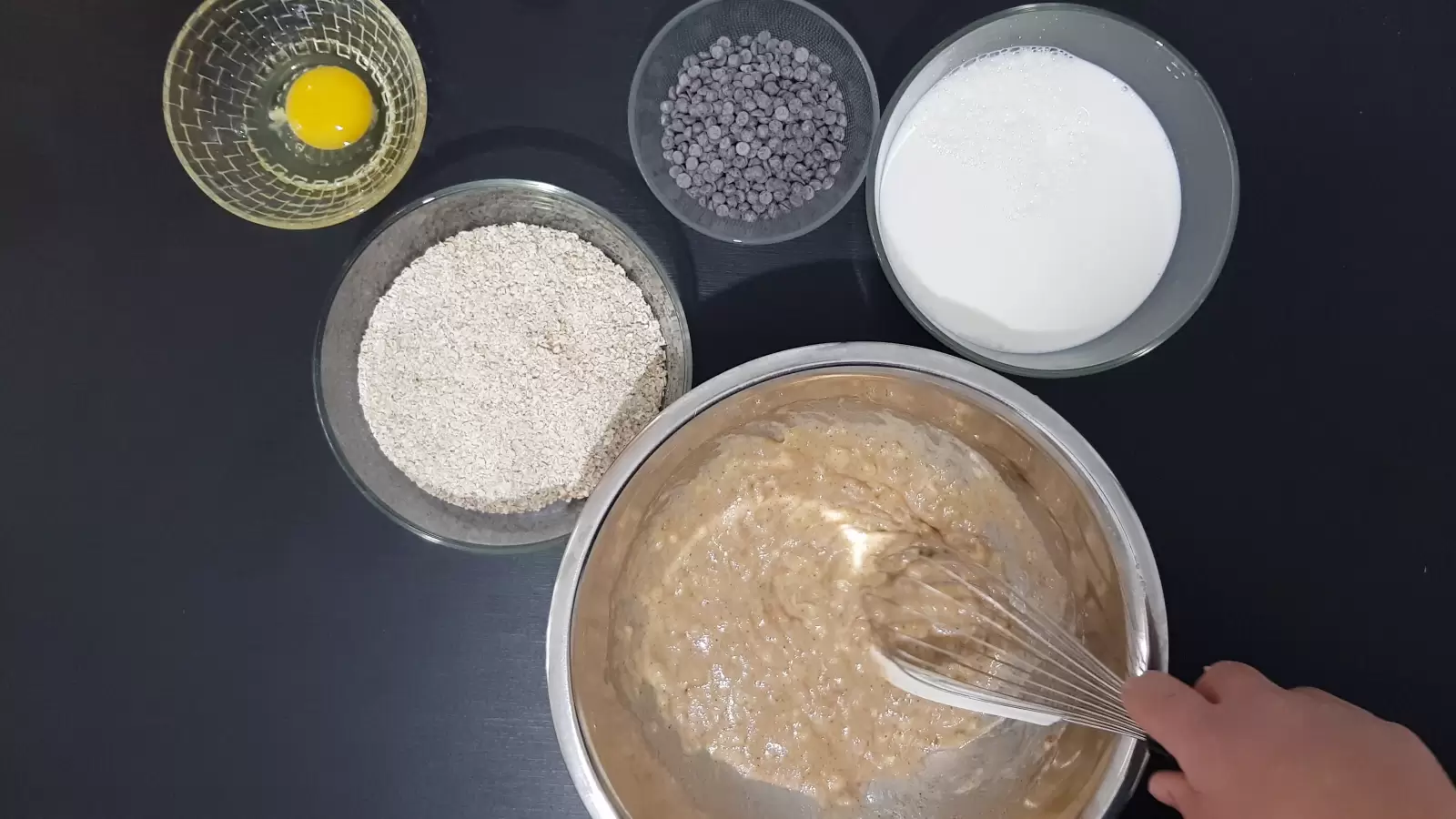 Bowl with chocolate chips, bowl with egg, bowl with oats, bowl with milk, whisk, mixture in a bowl