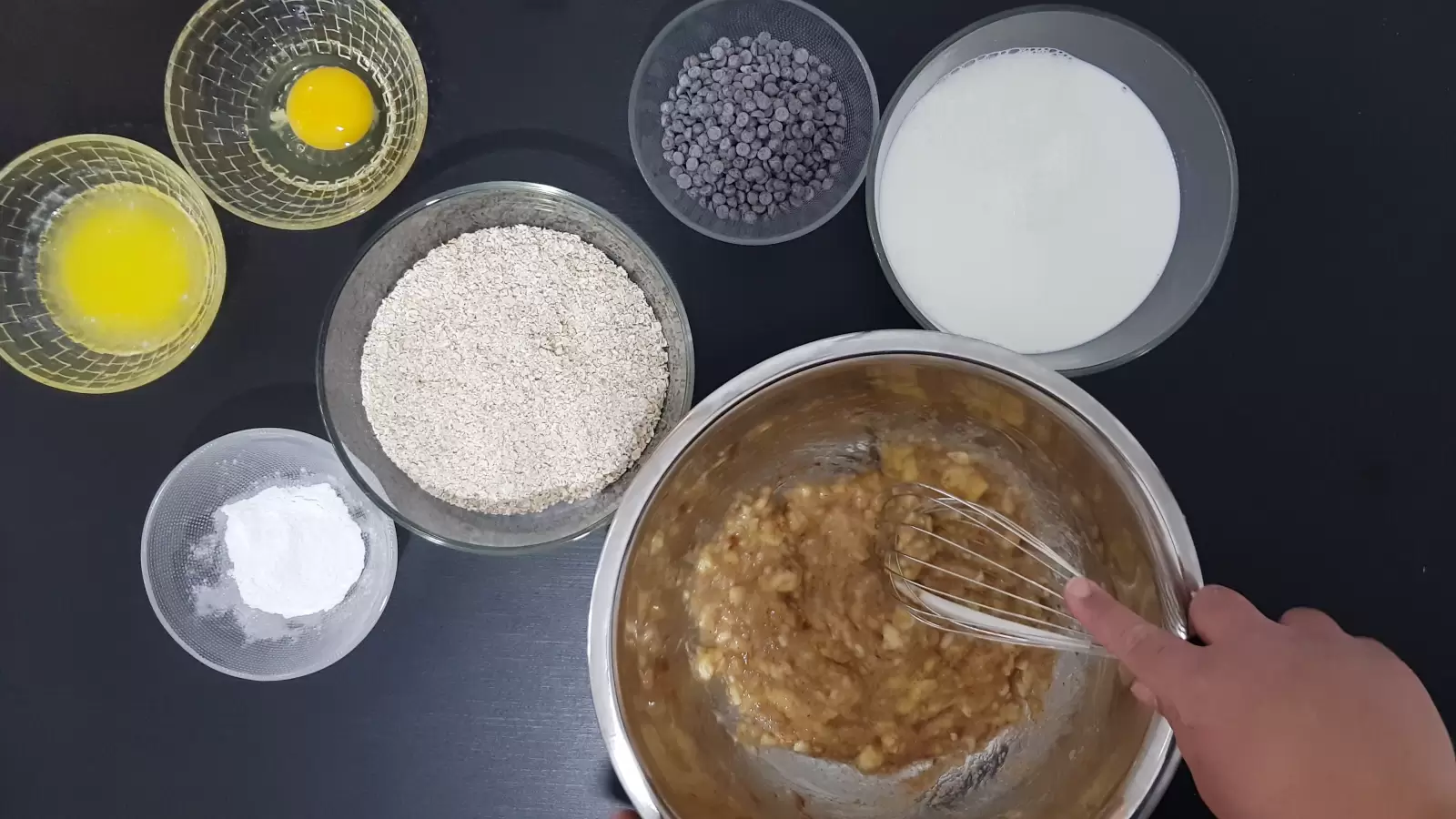 Bowl with baking powder, bowl with chocolate chips, bowl with egg, bowl with butter, bowl with oats, bowl with milk, whisk, mashed bananas in a bowl