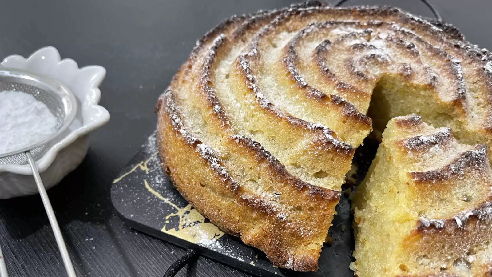 Orange and almond cake on a tray a slice of cake cut on the side there is a bowl with powdered sugar