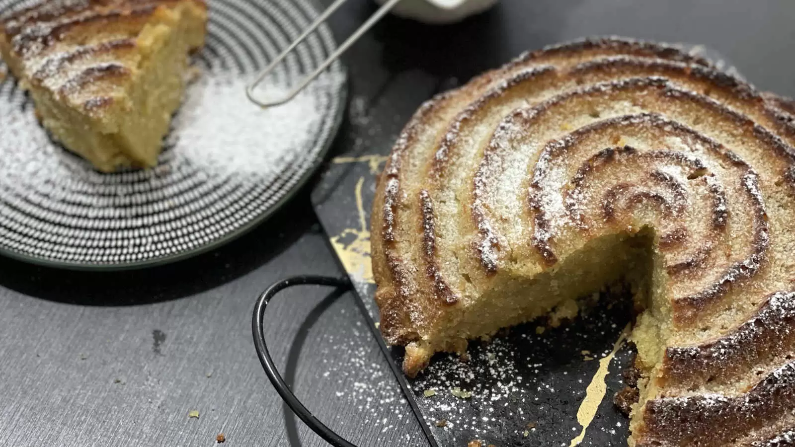 Orange and almond cake dish on a tray a slice of cake cut on the side there is a bowl with powdered sugar