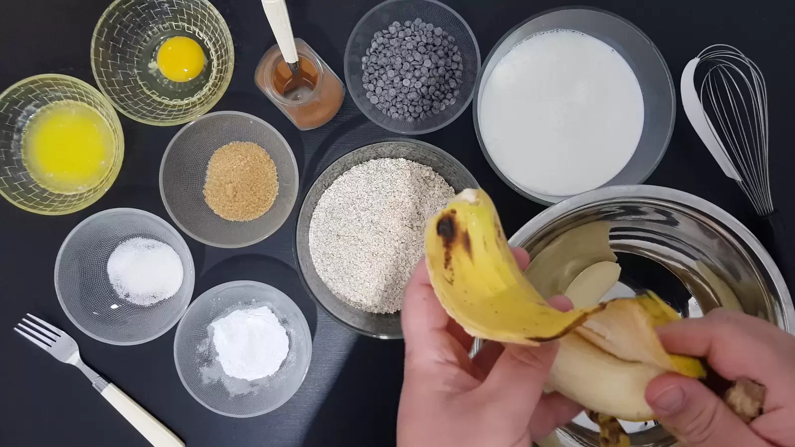 Bowl with baking powder, bowl with chocolate chips, cinnamon and spoon, bowl with egg, bowl with butter, bowl with brown sugar, bowl with oats, bowl with milk, bowl with vanilla sugar, bowl with baking powder, fork, whisk , a peeled banana over a bowl