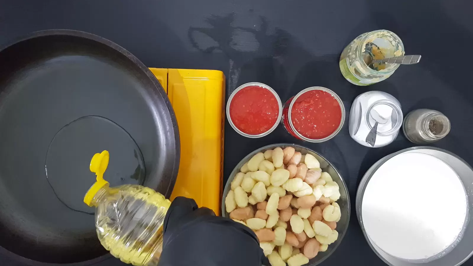 Crushed garlic with a spoon, a jar of pepper, a jar of salt with a spoon, 2 cans of crushed tomatoes, a bowl of whipping cream, a bowl of gnocchi, oil poured into a frying pan