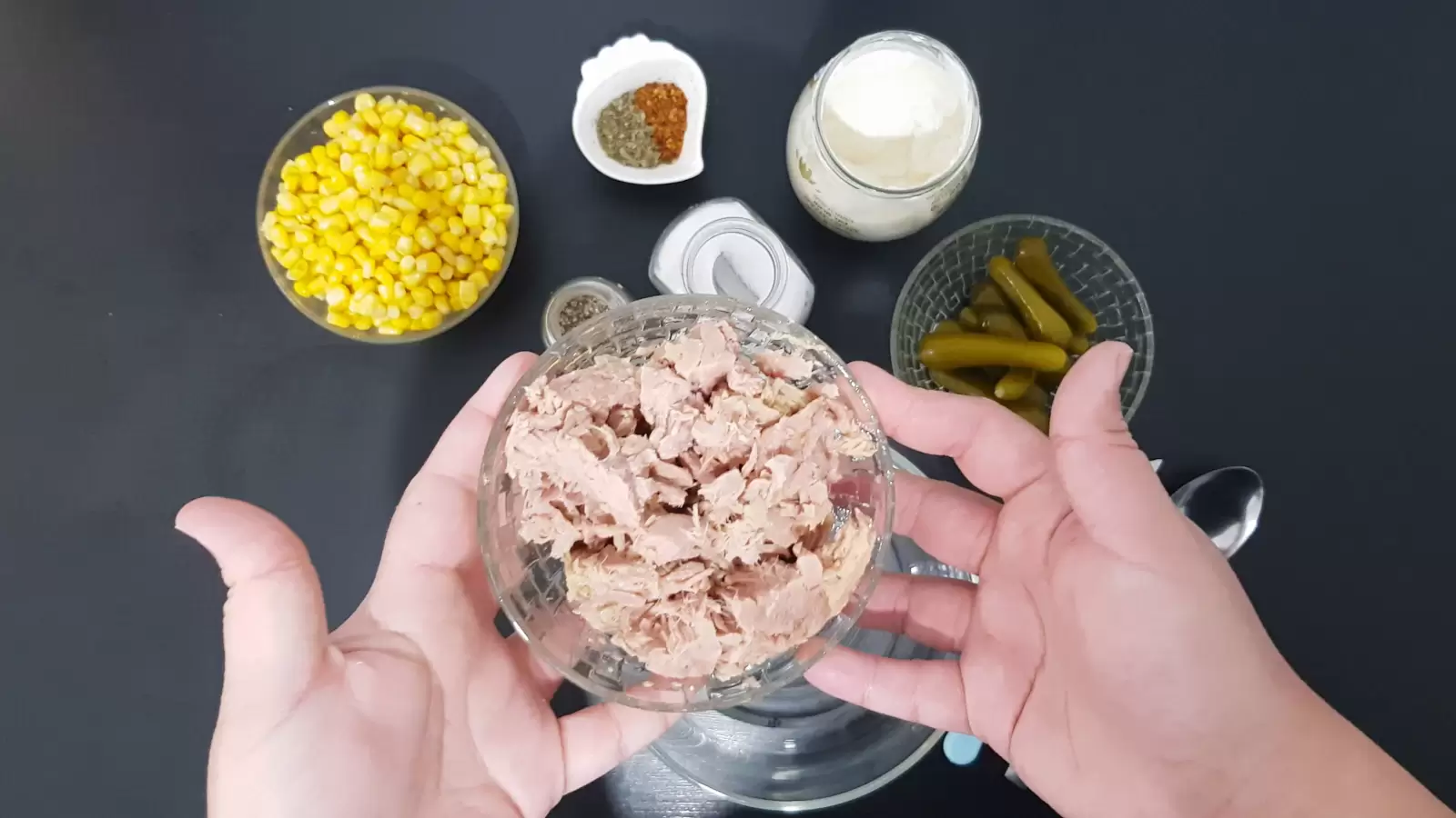 Bowl with tuna, bowl with corn, jar of salt, jar of mayonnaise, spoon, bowl with chili, bowl with pickles