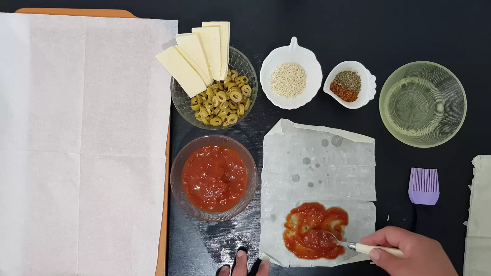 Bowl with oil, bowl with spices, bowl with sesame seeds, bowl with olives and yellow cheese, bowl with tomato sauce, filo dough with tomato sauce, brush, spoon and baking sheet with baking paper