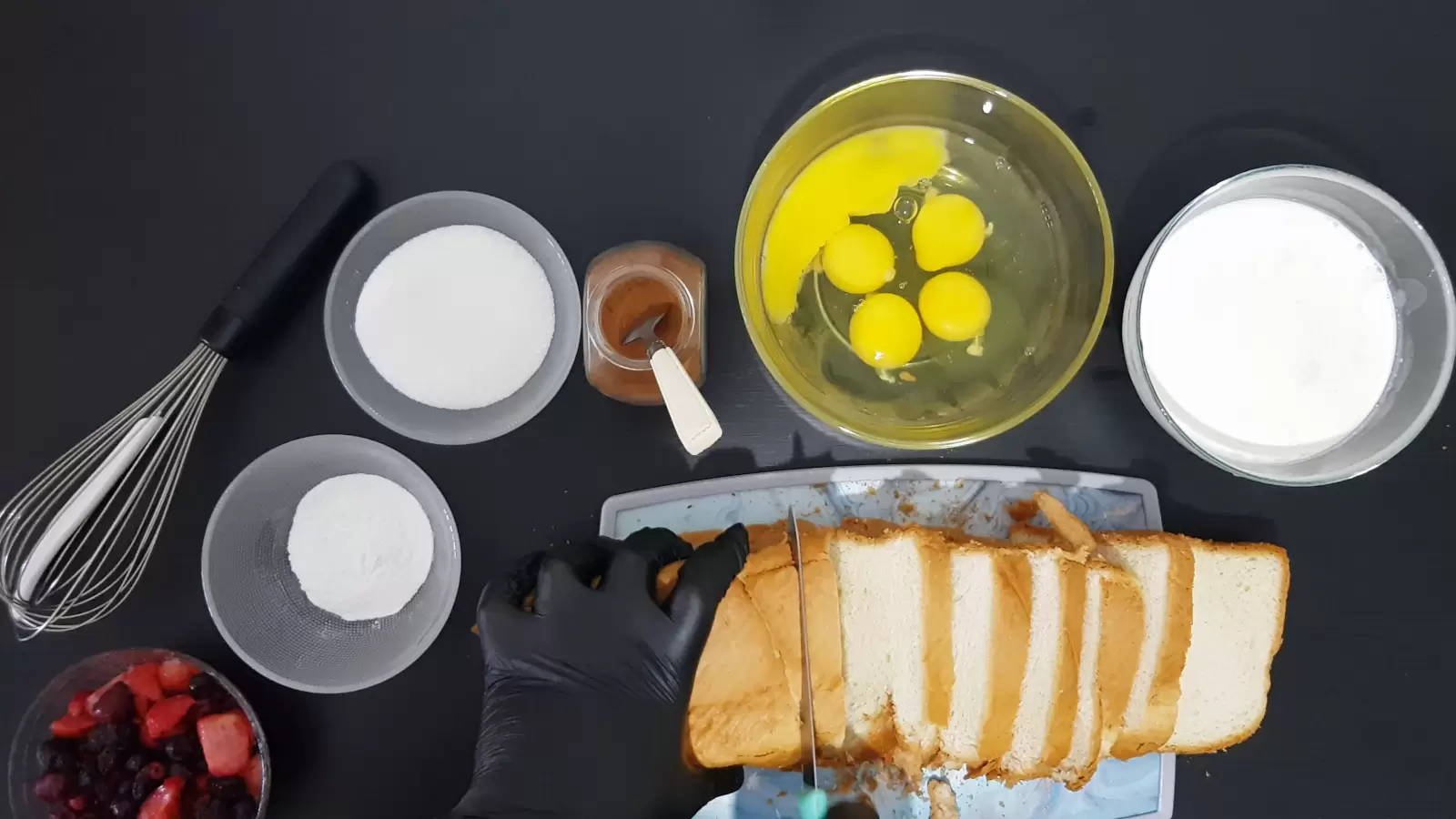 Bowl of heavy cream, bowl of eggs, jar of cinnamon spice and spoon, bowl of sugar, bowl of vanilla sugar, bowl of berries, sliced ​​bread on cutting board and knife, whisk