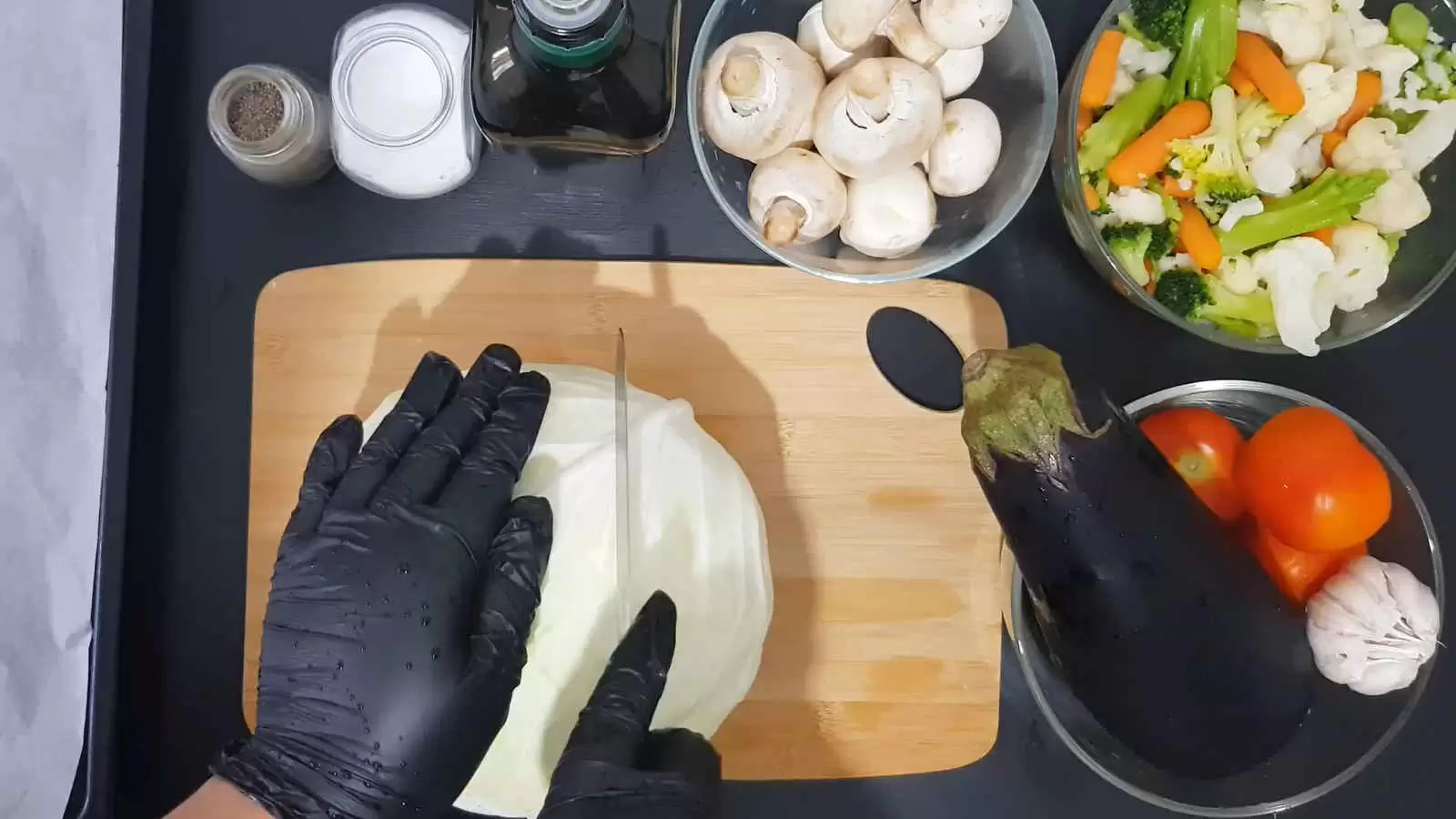 A bowl with cauliflower, carrots and broccoli, a bowl with mushrooms, a bottle of olive oil, a jar of salt, a jar of pepper, a bowl with garlic, tomatoes and eggplant. Cutting board with cabbage and knife