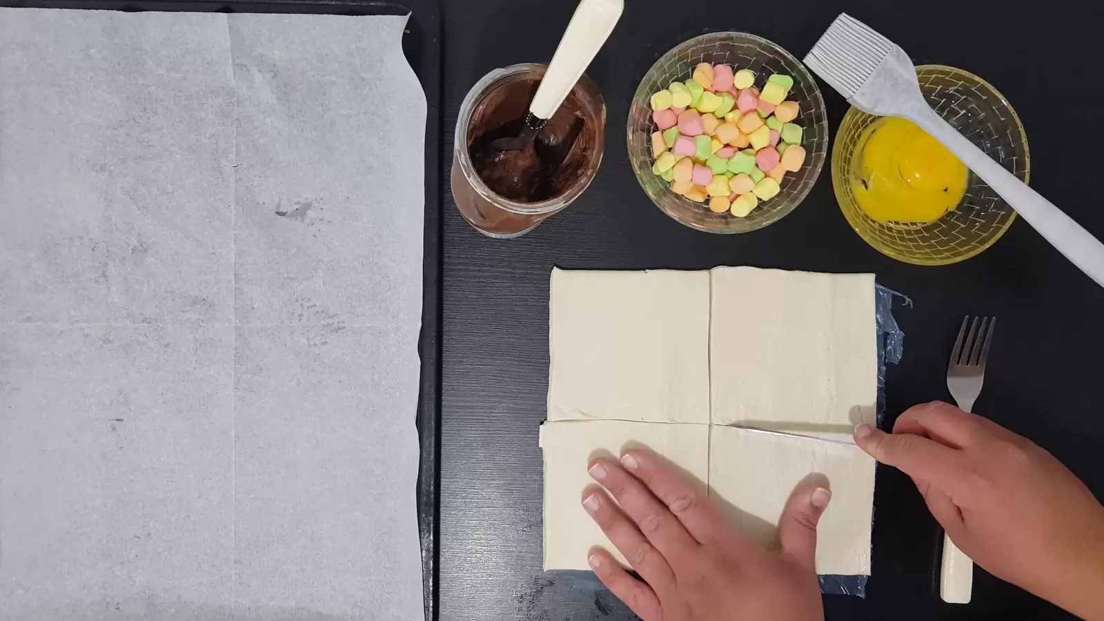 Bowl with egg and brush for spreading, bowl with marshmallows, chocolate spread, baking sheet with baking paper, puff pastry, knife and fork