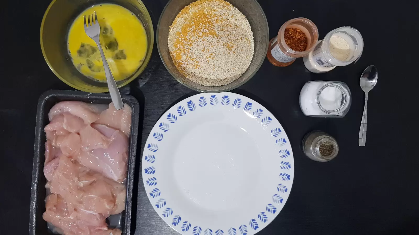 A bowl with eggs and a fork, a bowl with sesame seeds and breadcrumbs, a jar of chili, a jar of garlic powder, a jar of salt, a jar of pepper, a spoon, a plate, a mold with sliced ​​chicken breast