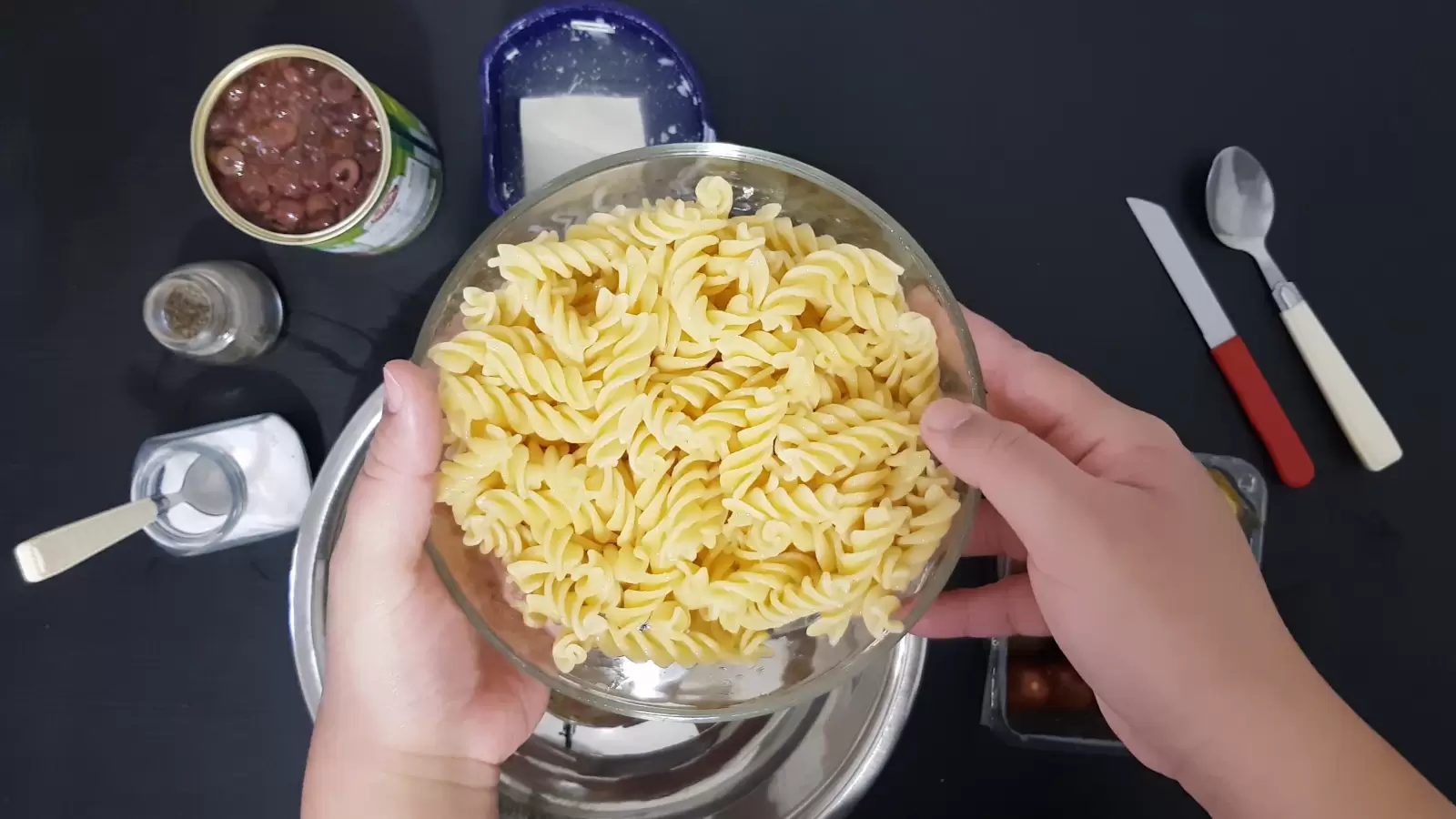 Spoon, knife, bowl with pasta, box with Bulgarian cheese, can with olives, jar of salt, jar of pepper