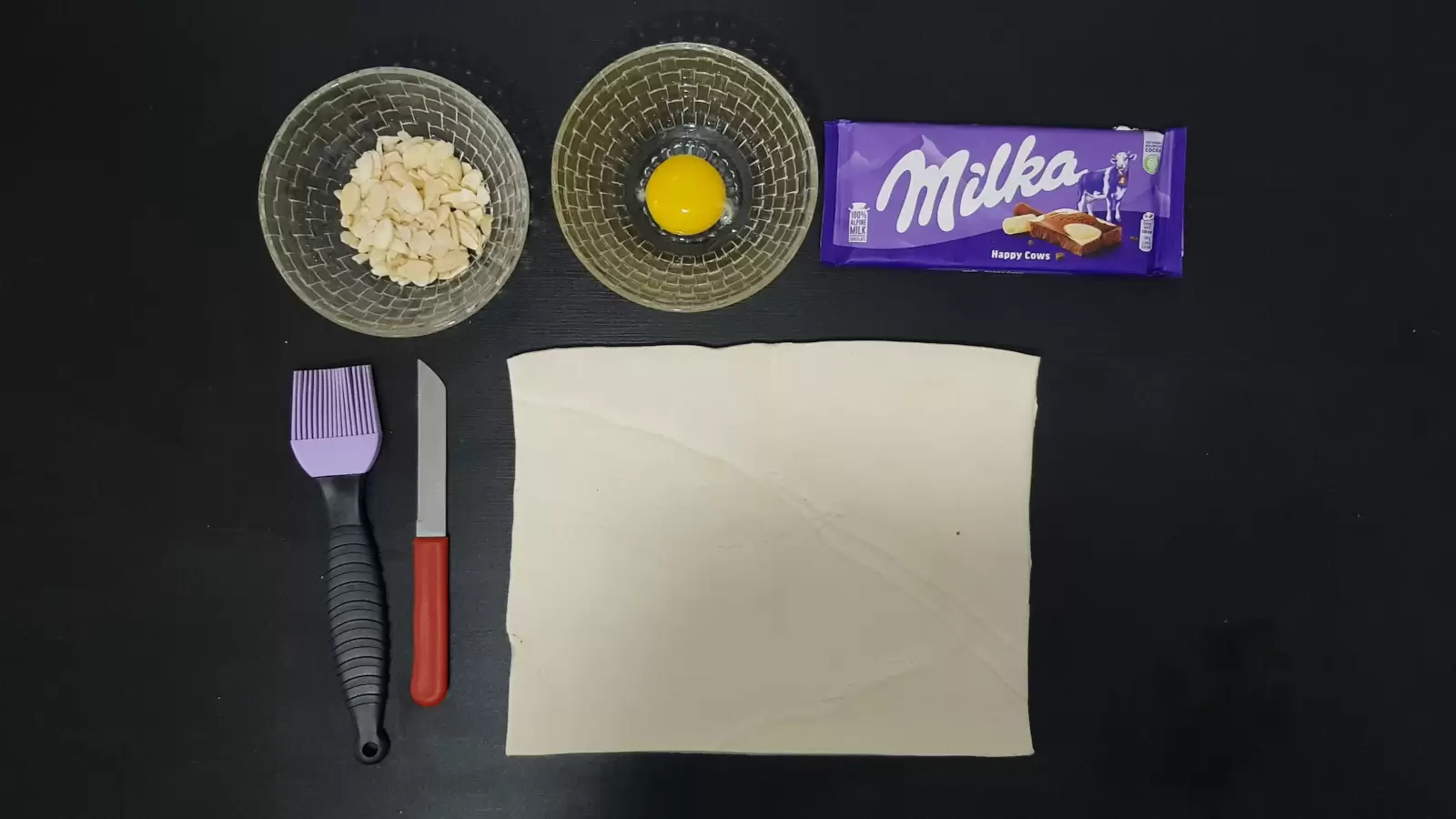 Milka chocolate bar, bowl with egg, bowl with almonds, puff pastry, knife, brush for spreading