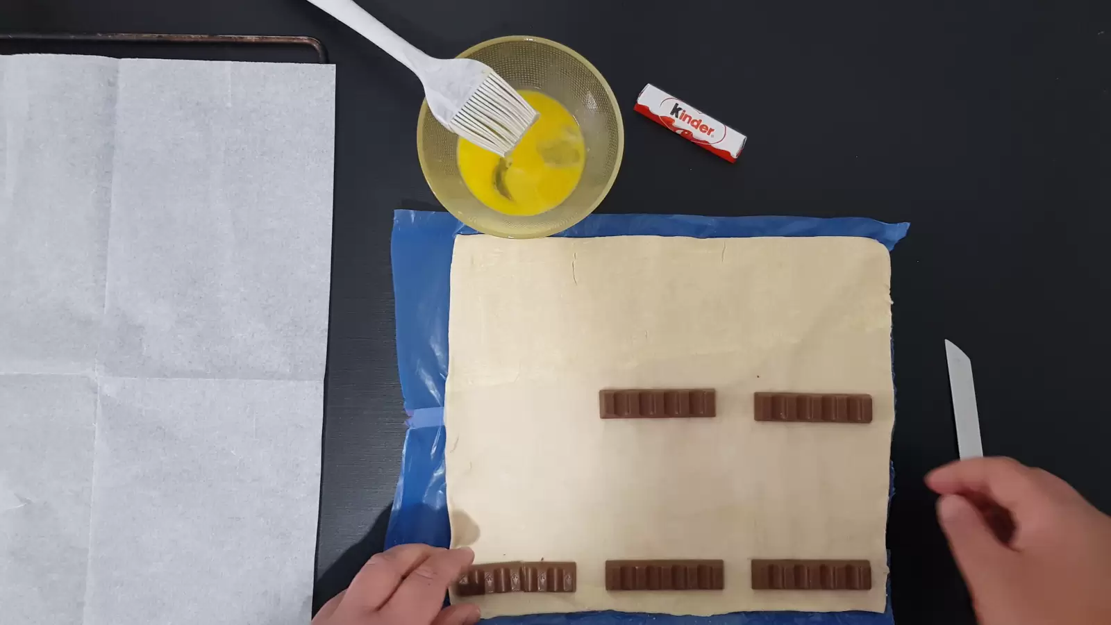 Six Kinder Buenos, puff pastry, a bowl with an egg and a brush, baking paper in an oven tray, a knife