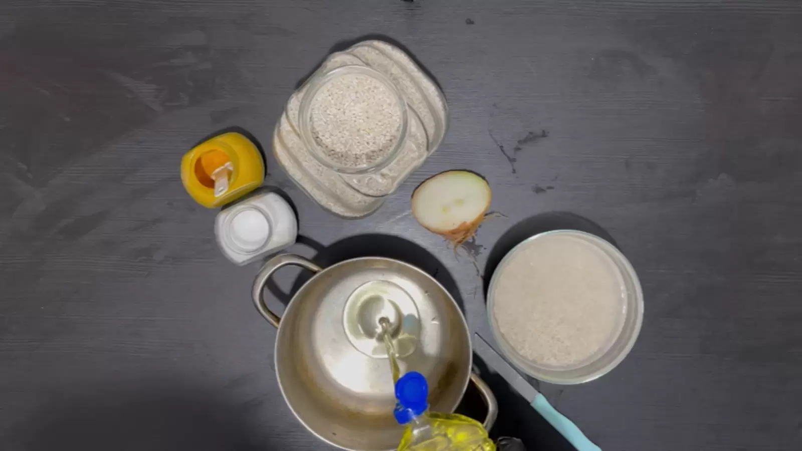 A jar of Pearl Barley Soup, half a medium onion, a bowl of Pearl Barley Soup and water, a pot with oil poured into it, a jar of salt, a jar of turmeric.