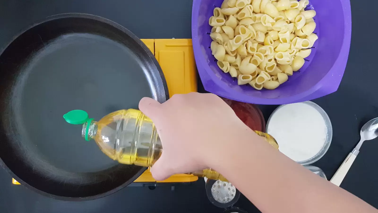 A bottle of oil, a frying pan, a bowl with pasta, a bowl with whipped cream, a spoon