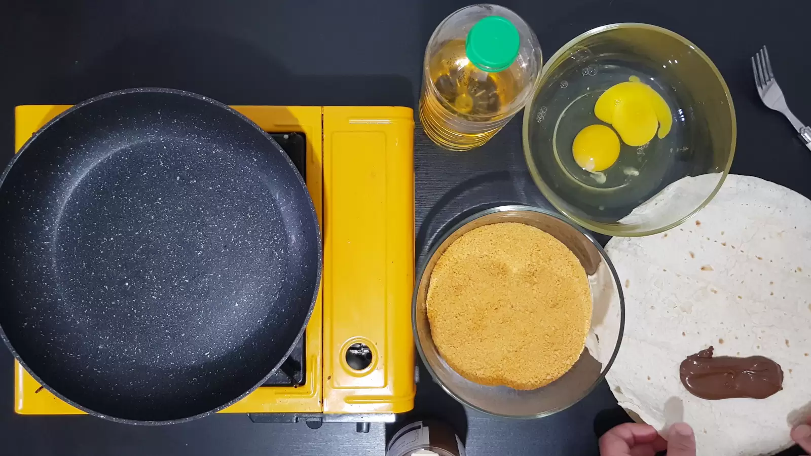 Bowl with egg, bottle of oil, bowl with breadcrumbs, tortilla and chocolate, stove and frying pan