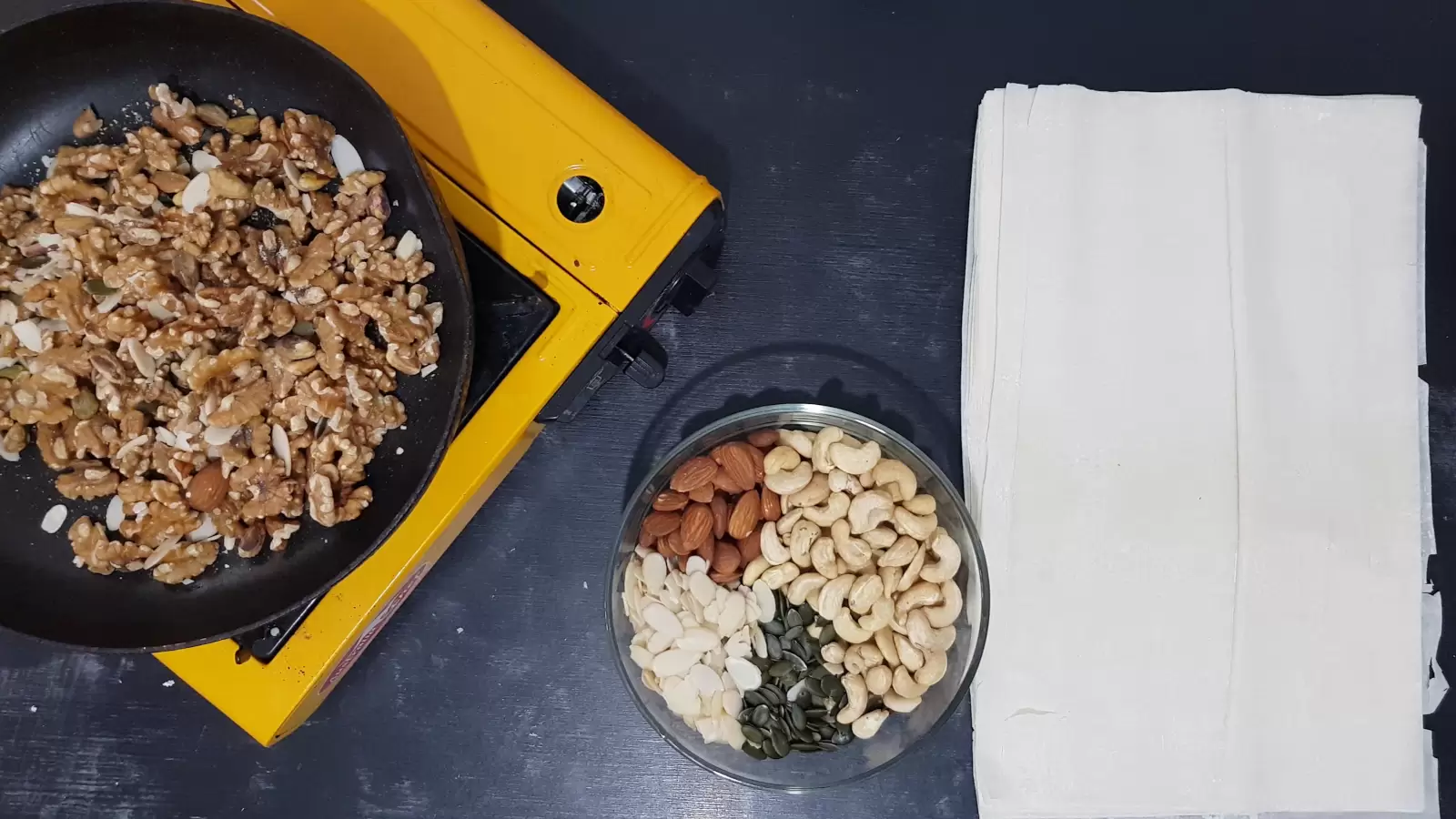 Filo sheets on a table, an assortment of nuts in a bowl, a pan with walnuts