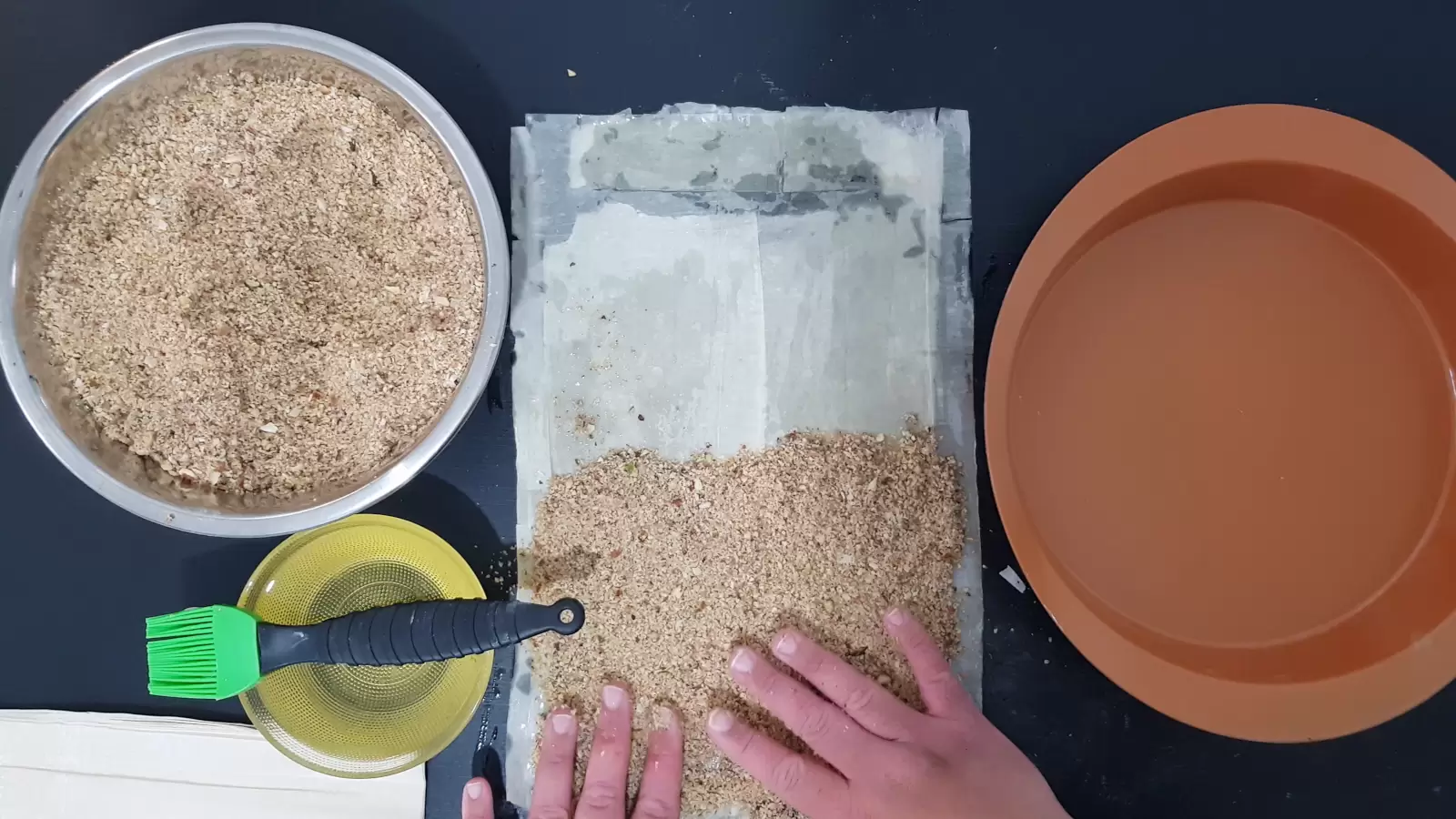 An oven tray, sheets of phyllo with the nut mixture on it, a bowl with oil and a brush for spreading, a bowl with a mixture of chopped nuts